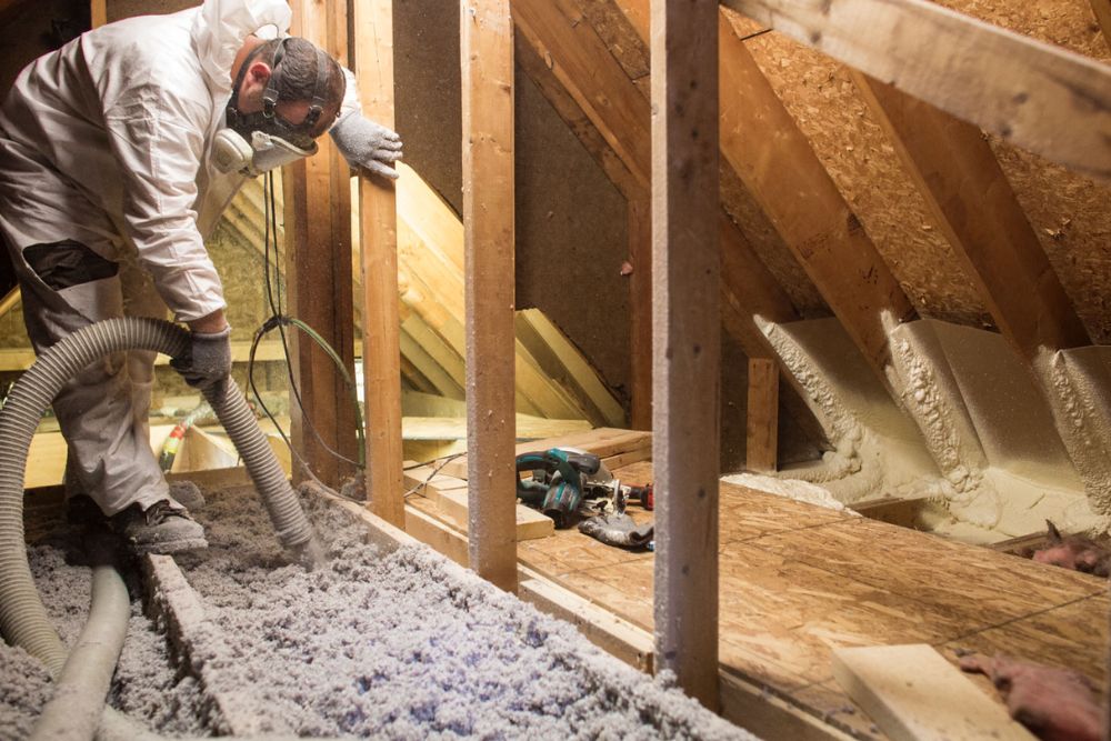 A man in white overalls installs insulation in an attic, surrounded by wooden beams and insulation materials.