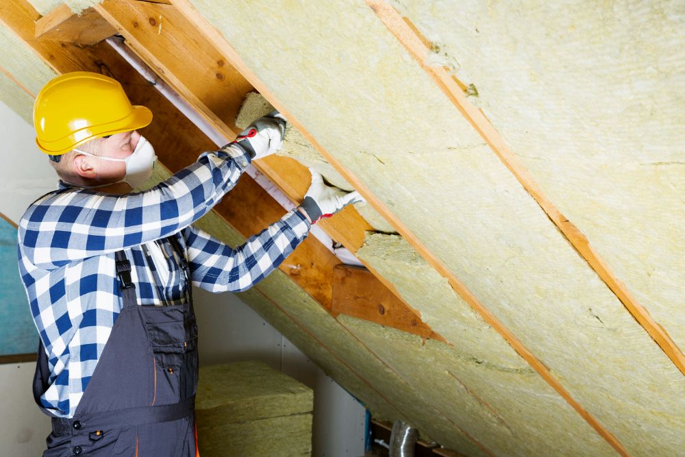 A man in a hard hat and overalls is repairing the roof in an attic, focused on his work amidst wooden beams.