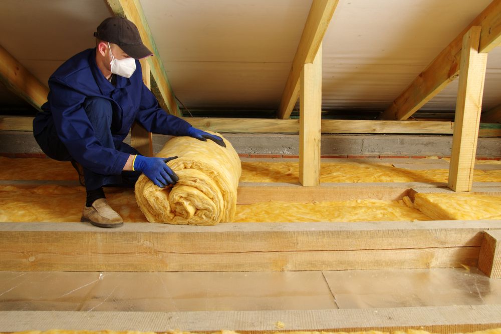 A worker in a mask rolls out insulation material in an attic, surrounded by wooden beams and yellow insulation batts.