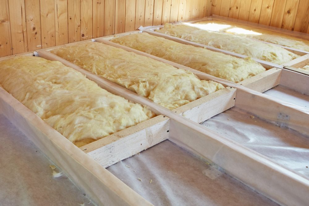 Insulation batts installed between wooden framing in a bright, unfinished room with wooden walls.