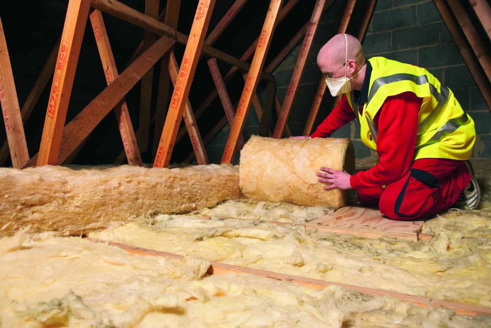 A worker in a safety vest installs insulation in an attic, wearing a mask while handling insulation material.